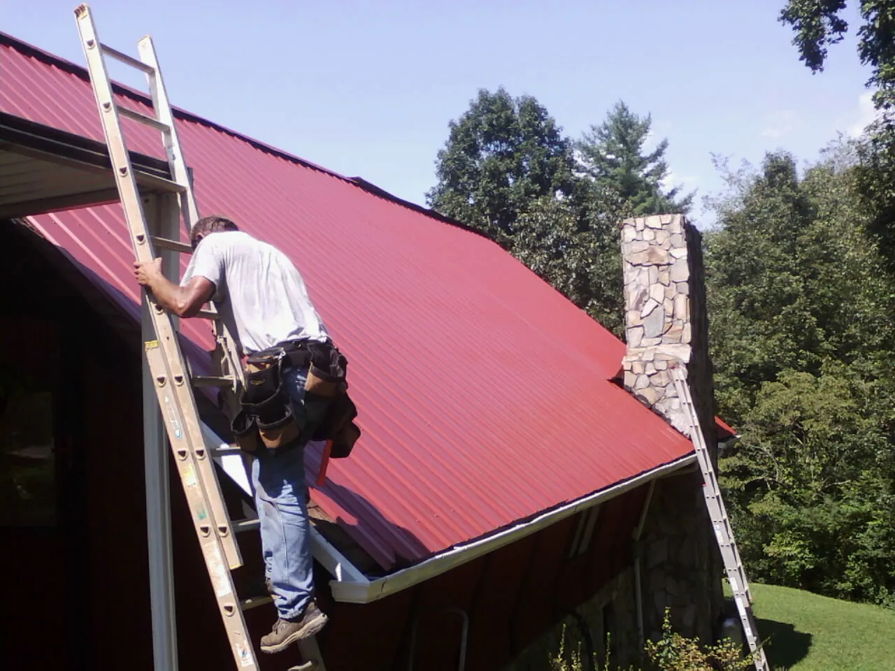 Roofer on a ladder beside a red metal roof and stone chimney — Soffit & Fascia Repair by Roofing Brothers in Annawan